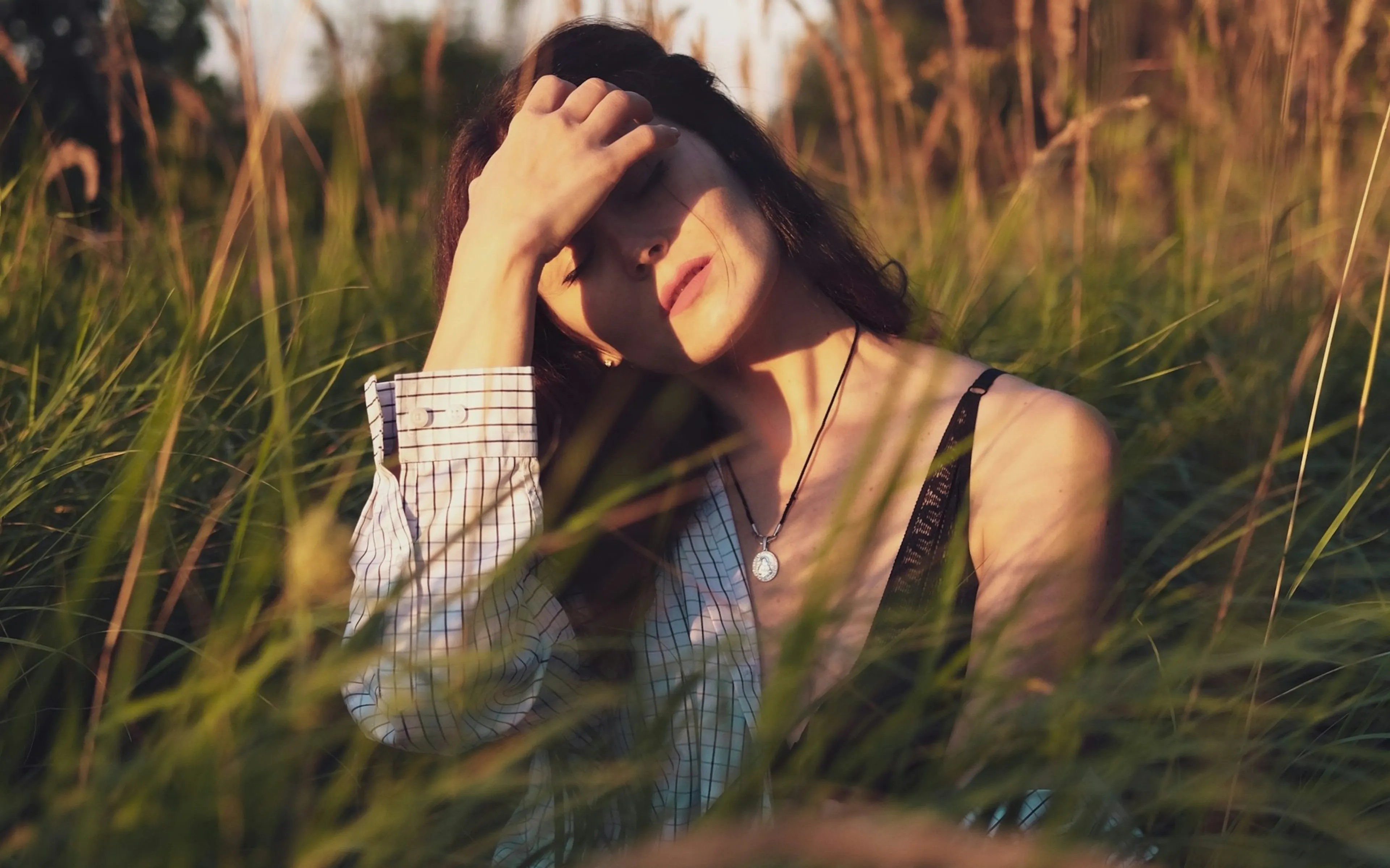 Een vrouwenportret van een jonge vrouw in een open veld, gefotografeerd in warm zonlicht. De scène benadrukt haar sombere houding en gezichtsuitdrukking terwijl ze een hand tegen haar voorhoofd houdt.