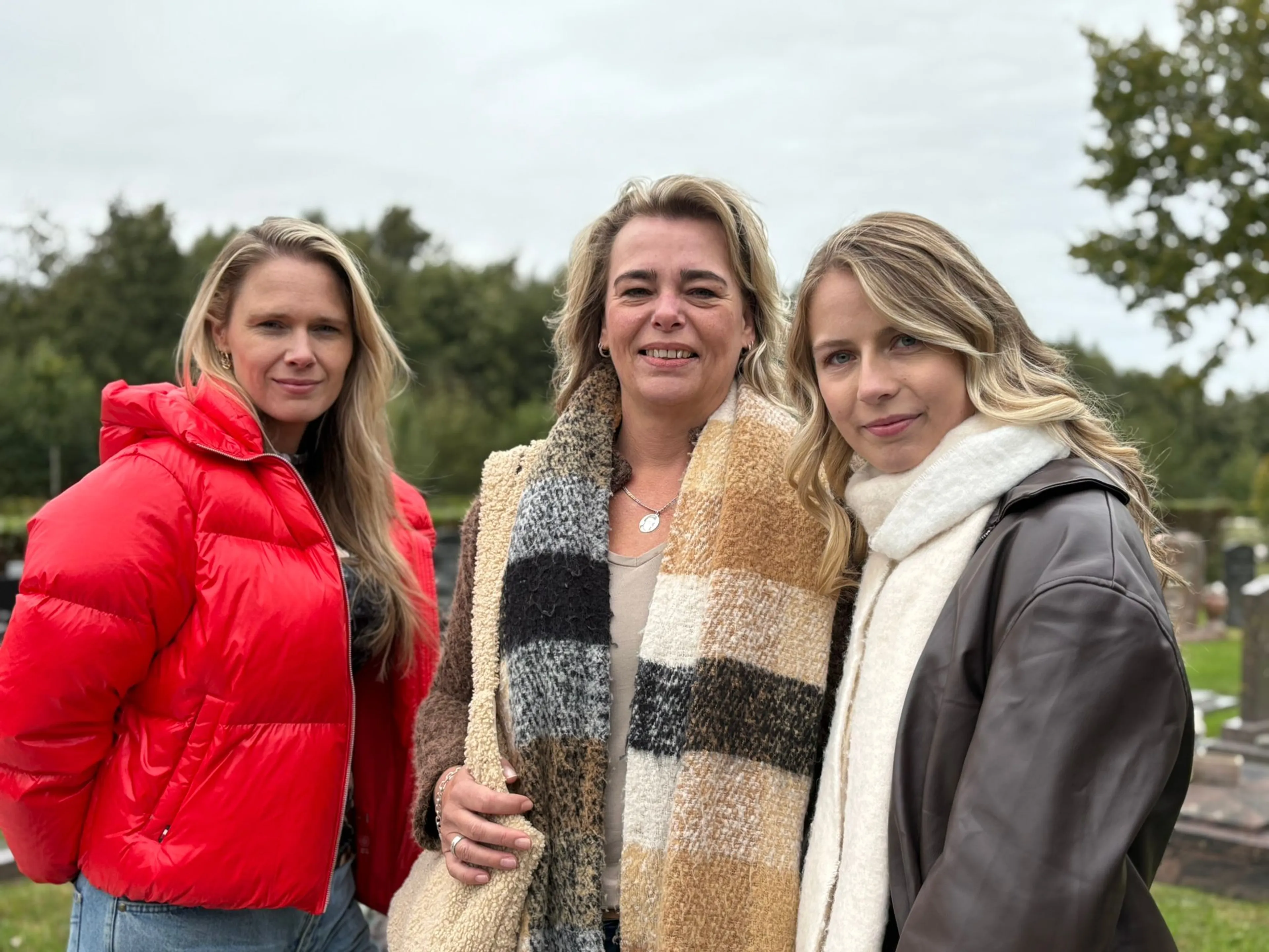 Begraafplaats met drie vrouwen: een gezelschap staand buiten in jasjes en sjaals, terwijl de achtergrond een groen parkachtige terrein met grafstenen en een grijze, bewolkte lucht toont. De beschrijving benadrukt de nabijheid en expressies van de drie vrouwen zonder verdere interpretatie.