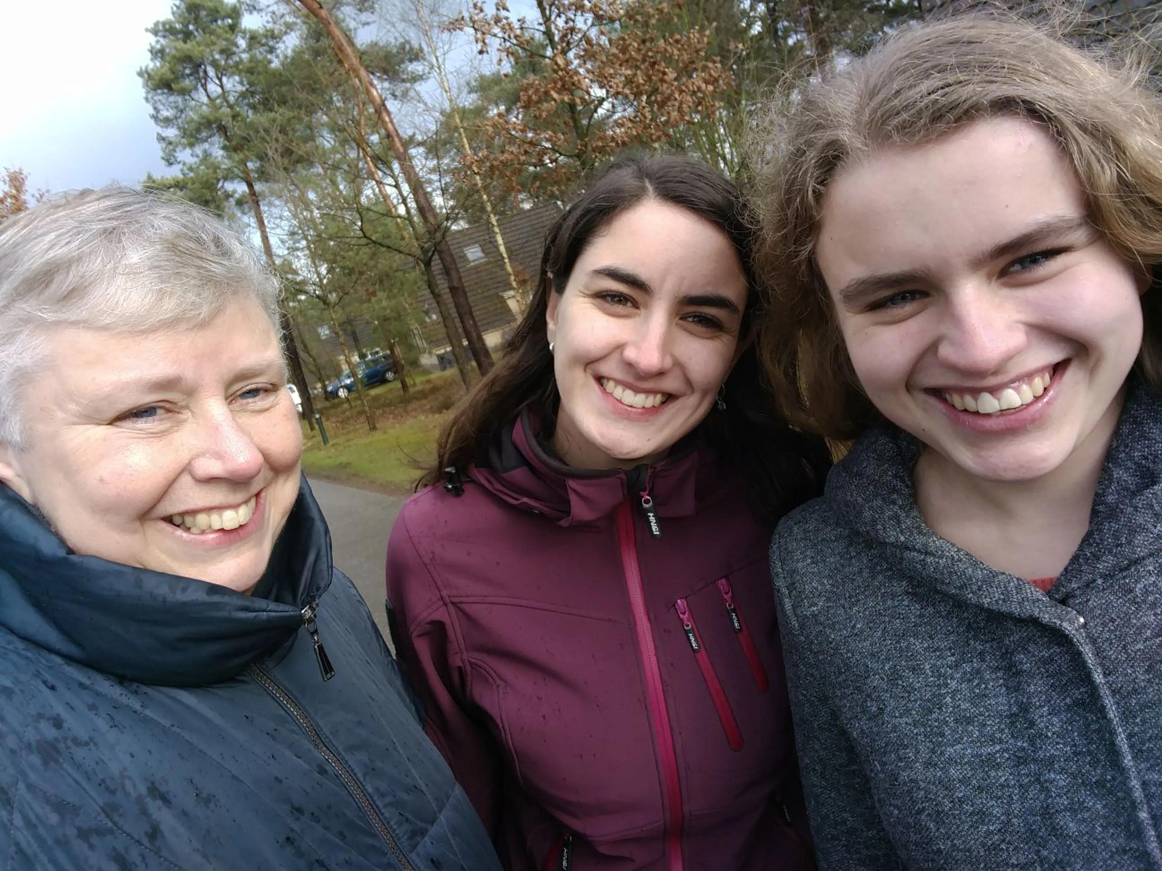 Drie personen poseren voor een close-up selfie buiten, terwijl ze breed lachen naar de camera. Op de achtergrond is een natte, bewolkte dag te zien met hoge bomen, een gebouw en geparkeerde auto's, wat een rustige woon- of parkachtige omgeving suggereert.