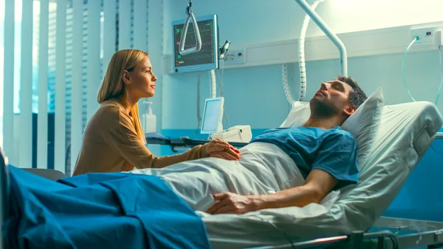 Een vrouw kijkt gelaten naar een man die in een ziekenhuisbed ligt met gesloten ogen.
